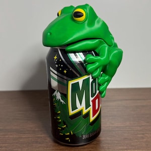 May include: A green frog-shaped can holder perched on a Mountain Dew Zero Sugar can. The can is black with green and yellow accents, and the frog has yellow eyes. The can holder adds a fun, unique touch to any beverage.