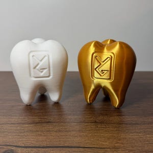 May include: Two three-dimensional tooth-shaped figurines, one matte white and one metallic gold. Each figurine features a square embossed logo on the front containing the stylized letters YG. The objects have a smooth, layered texture.
