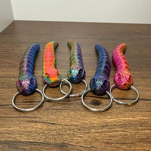 May include: Five colorful snake-shaped keychains. Each keychain features a different color scheme, including blue, rainbow, green, dark blue, and pink. The keychains have a metallic sheen and are attached to silver key rings. The keychains are displayed on a wooden surface.