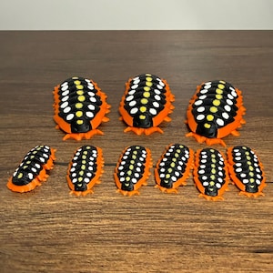 May include: A collection of ten orange, black, and white insect-like figurines. The figurines have a segmented body design with a row of white dots down the center. The figurines are arranged on a brown wooden surface.