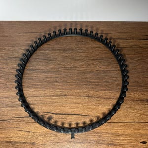 May include: A black circular knitting loom with evenly spaced pegs. The loom is made of plastic and sits on a wooden surface. The pegs are designed to hold yarn for knitting projects. The loom is a crafting tool.