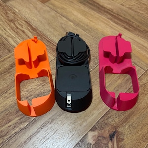 May include: Three brightly colored charging station holders. An orange holder is on the left, a black charging station with a cord is in the center, and a pink holder is on the right. The holders are made of plastic and have a unique design.