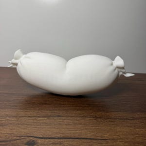 May include: A solid white, elongated, cylindrical object with a smooth, matte finish. The object features pinched, tied-off ends that resemble the twisted casing of a sausage, creating a distinct, symmetrical, and curved shape in a neutral tone.