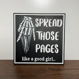 May include: Black and white square sign with the text "SPREAD THOSE PAGES" and "like a good girl.." in white lettering. A skeleton hand graphic is on the left side of the sign.