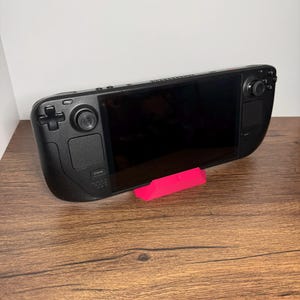 May include: A black handheld gaming console with a large screen, buttons, and joysticks. A bright pink stand supports the console on a wooden surface. The console has the word "STEAM" printed on it.