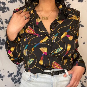 May include: Black button-down blouse with a gold rope and tassel pattern. The blouse features long sleeves and a collared neckline. The tassels are in shades of yellow, pink, blue, and green. A black belt and light wash jeans complete the look.