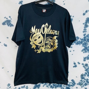 Pode incluir: Uma camiseta preta com um gráfico dourado das palavras "New Orleans" e duas máscaras teatrais. As máscaras são uma cara feliz e uma cara triste.