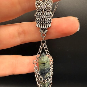 May include: Silver-toned owl pendant necklace with a green and black crystal. The owl pendant has detailed feather and eye designs. The crystal is encased in a silver chain. The necklace is displayed against a dark background.