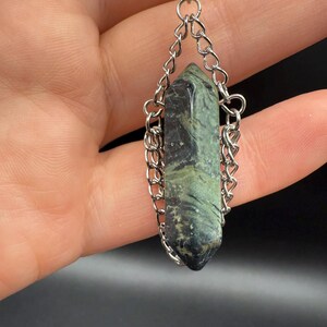 May include: A pendant featuring a green and black crystal suspended by a silver chain. The crystal is a long, pointed shape with natural variations in color. The chain is made of small, interlocking links, and the pendant's top has a decorative silver cap.