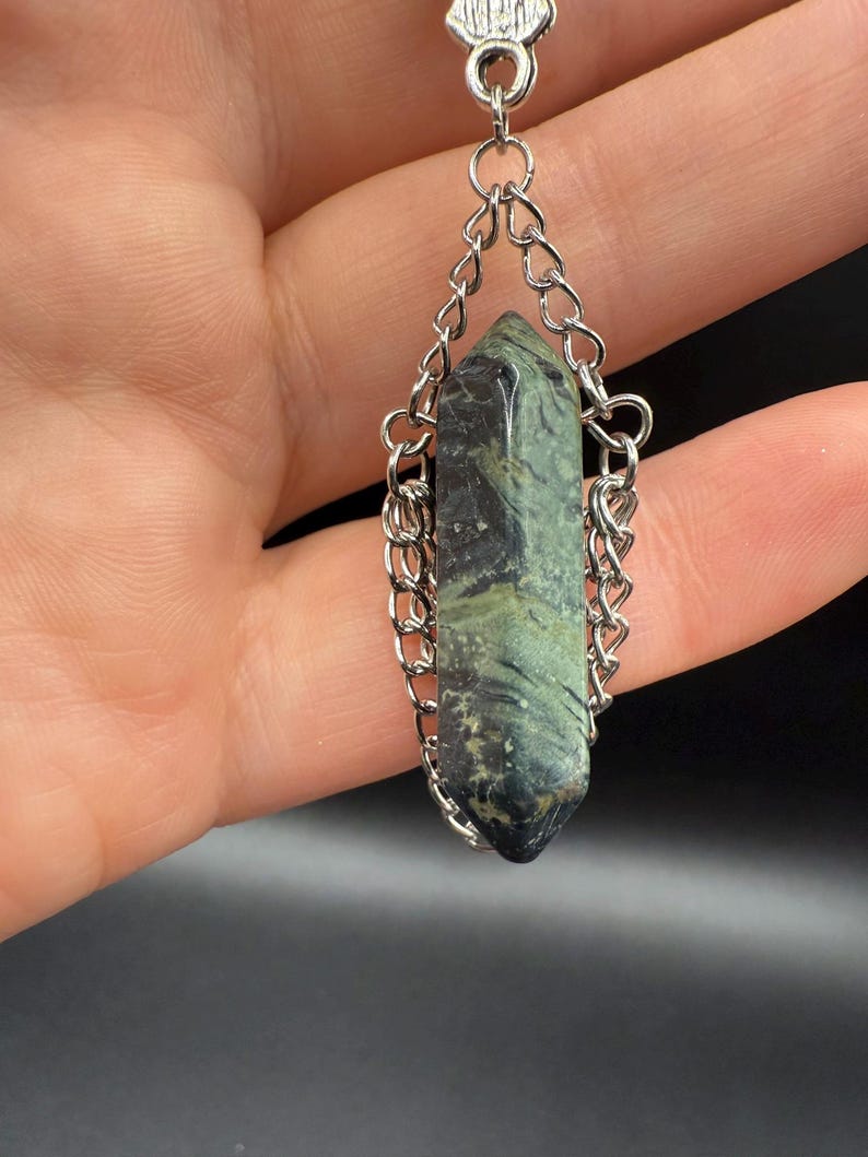 May include: A pendant featuring a green and black crystal suspended by a silver chain. The crystal is a long, pointed shape with natural variations in color. The chain is made of small, interlocking links, and the pendant's top has a decorative silver cap.