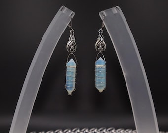 May include: A pair of earrings featuring light blue crystal pendants wrapped in silver wire. The earrings have ornate silver accents at the top and are displayed on clear stands against a dark background. The crystals are approximately 1.5 inches long.