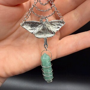 May include: Silver-toned pendant necklace featuring a detailed moth design with chain accents. A green crystal is wire-wrapped and suspended below the moth. The necklace is held in a hand against a dark background.