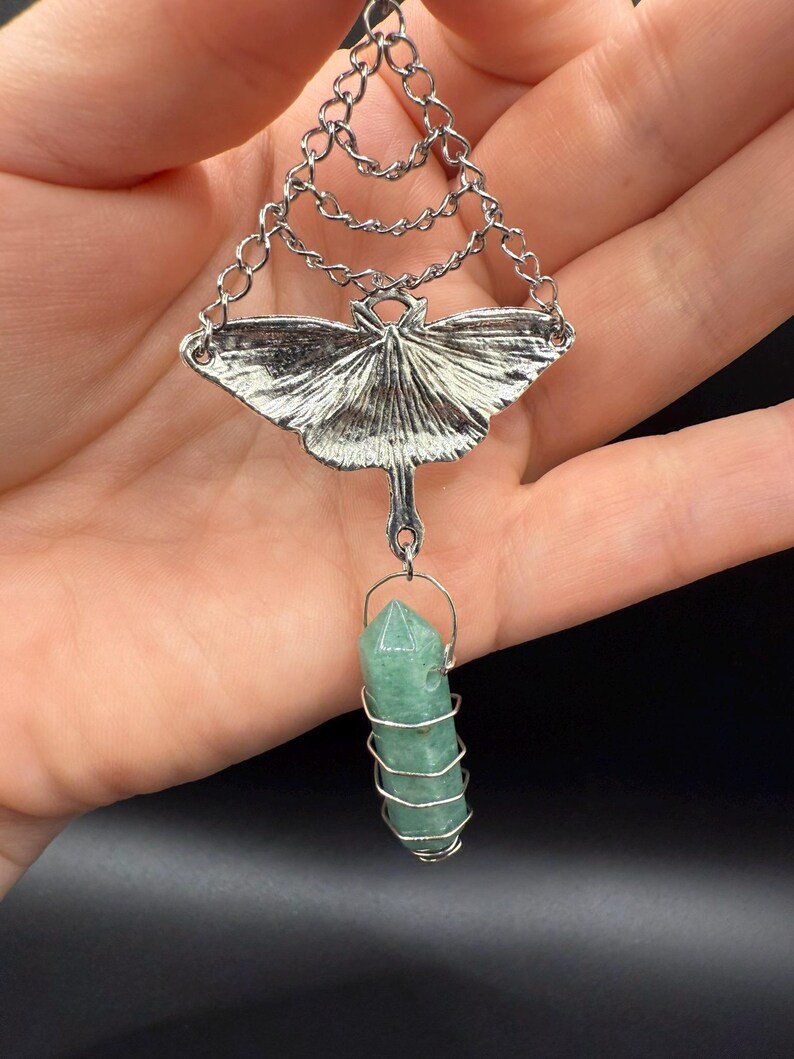 May include: Silver-toned pendant necklace featuring a detailed moth design with chain accents. A green crystal is wire-wrapped and suspended below the moth. The necklace is held in a hand against a dark background.