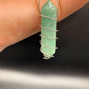 May include: A green crystal pendant with a silver wire wrap. The crystal is a light green color and is shaped like a long, six-sided prism. The silver wire is wrapped around the crystal in a spiral pattern, and a silver loop is at the top for hanging.