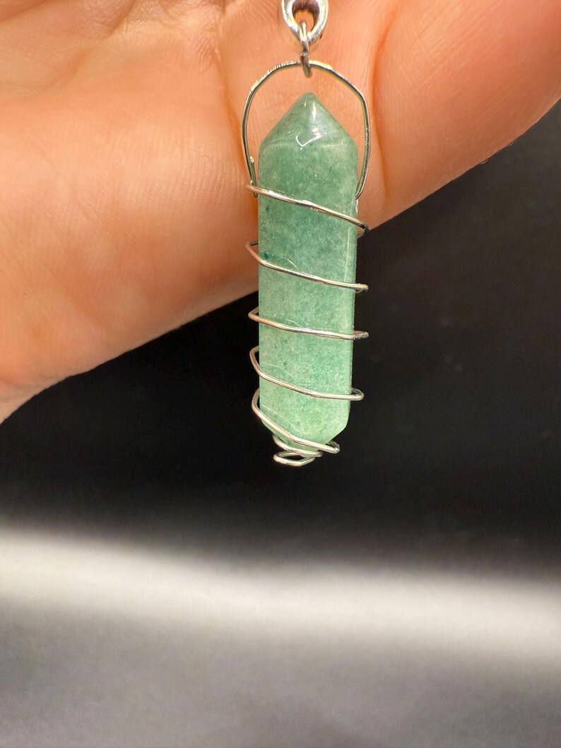 May include: A green crystal pendant with a silver wire wrap. The crystal is a light green color and is shaped like a long, six-sided prism. The silver wire is wrapped around the crystal in a spiral pattern, and a silver loop is at the top for hanging.
