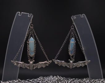 May include: A pair of silver-toned bat earrings. Each earring features a bat pendant with outstretched wings, suspended from a chain. A light blue crystal hangs below the chain. The earrings are displayed on clear stands against a dark background.