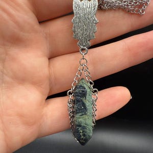 May include: Silver-toned pendant necklace featuring a textured owl-shaped top piece and a dark green and black crystal pendant. The crystal is encased in a silver chain. The necklace is displayed against a dark background.