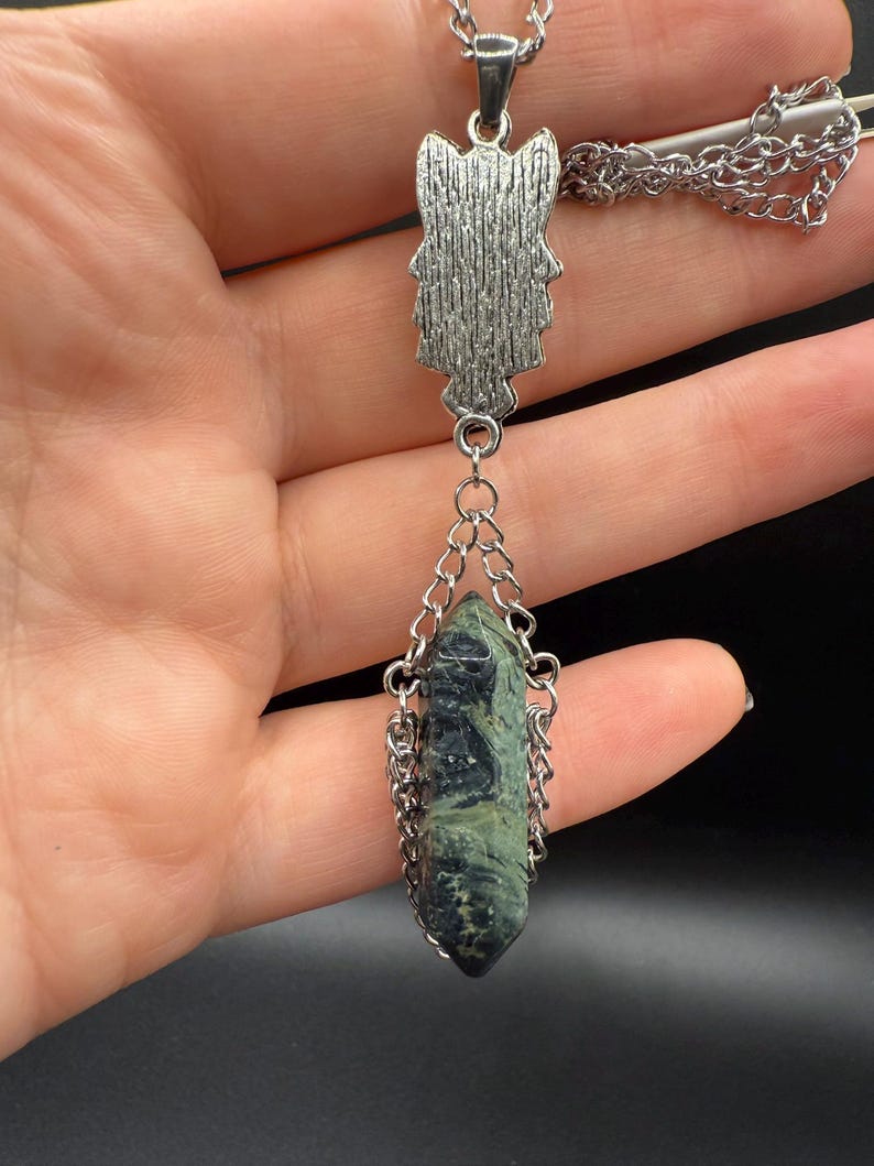 May include: Silver-toned pendant necklace featuring a textured owl-shaped top piece and a dark green and black crystal pendant. The crystal is encased in a silver chain. The necklace is displayed against a dark background.