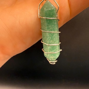 May include: A green aventurine crystal pendant wrapped in silver-colored wire. The crystal is a light green color and has a hexagonal shape. The wire is wrapped around the crystal in a spiral pattern, creating a unique and eye-catching design.