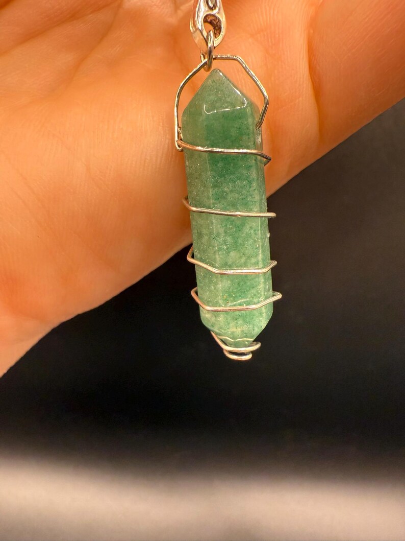 May include: A green aventurine crystal pendant wrapped in silver-colored wire. The crystal is a light green color and has a hexagonal shape. The wire is wrapped around the crystal in a spiral pattern, creating a unique and eye-catching design.