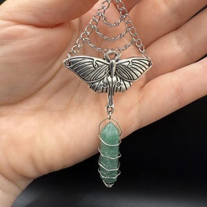 May include: Silver-toned pendant necklace featuring a detailed moth design with layered chain accents. A green crystal is suspended below, wrapped in silver wire. The pendant is a unique piece of jewelry.