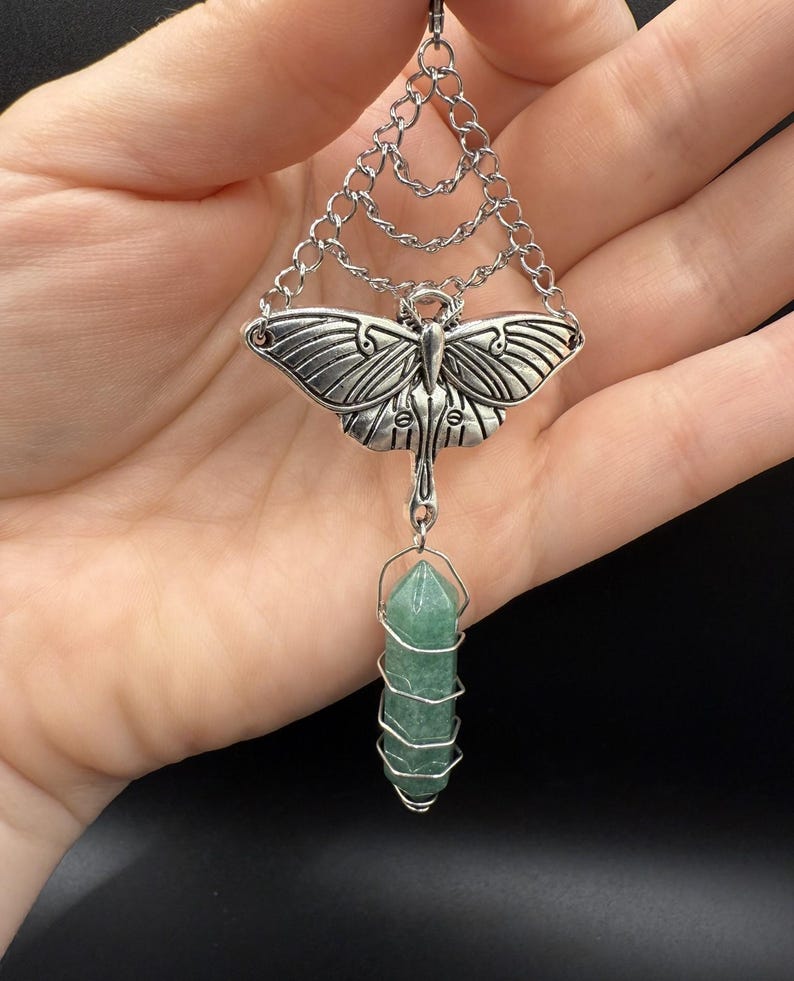 May include: Silver-toned pendant necklace featuring a detailed moth design with layered chain accents. A green crystal is suspended below, wrapped in silver wire. The pendant is a unique piece of jewelry.
