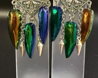 May include: A pair of dangle earrings featuring iridescent beetle wings in shades of green, blue, and gold. The earrings have ornate silver-toned diamond-shaped tops and are accented with silver chains and cone-shaped charms. The earrings are displayed on a clear plastic stand.