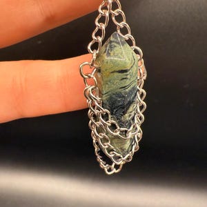 May include: A pendant featuring a green and black stone encased in a silver chain. The stone is a pointed, elongated shape. The chain is a delicate, linked design, creating a cage around the stone. The background is a dark, solid color.