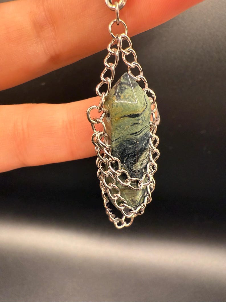 May include: A pendant featuring a green and black stone encased in a silver chain. The stone is a pointed, elongated shape. The chain is a delicate, linked design, creating a cage around the stone. The background is a dark, solid color.