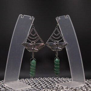 May include: Silver-toned earrings featuring a moth design with intricate wing details. The earrings have a chain design above the moth and a green crystal wrapped in silver wire. The earrings are displayed on clear stands against a dark background.
