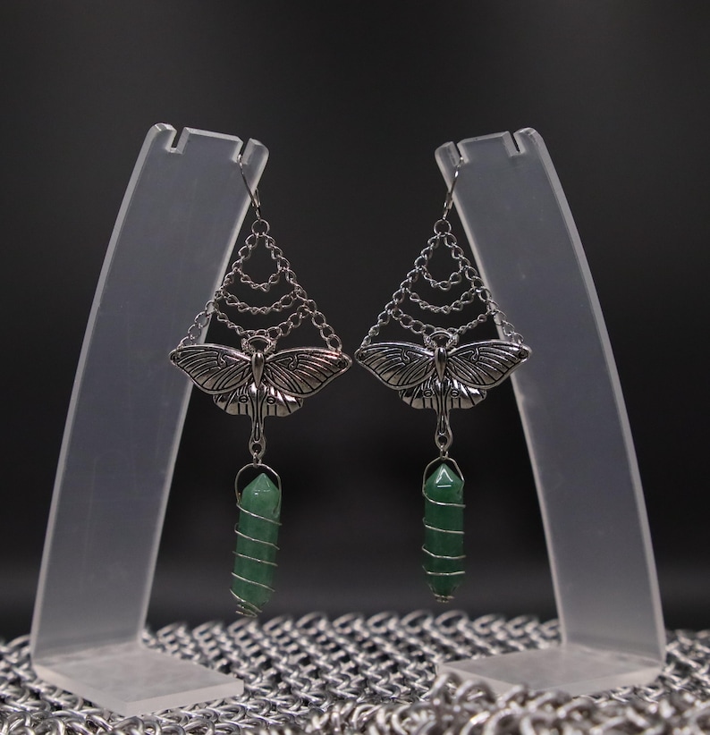 May include: Silver-toned earrings featuring a moth design with intricate wing details. The earrings have a chain design above the moth and a green crystal wrapped in silver wire. The earrings are displayed on clear stands against a dark background.