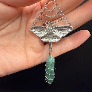 May include: A silver-toned pendant featuring a moth design with a green crystal. The moth has detailed wing patterns and hangs from a chain. The green crystal is wrapped in silver wire, creating a unique and elegant accessory.