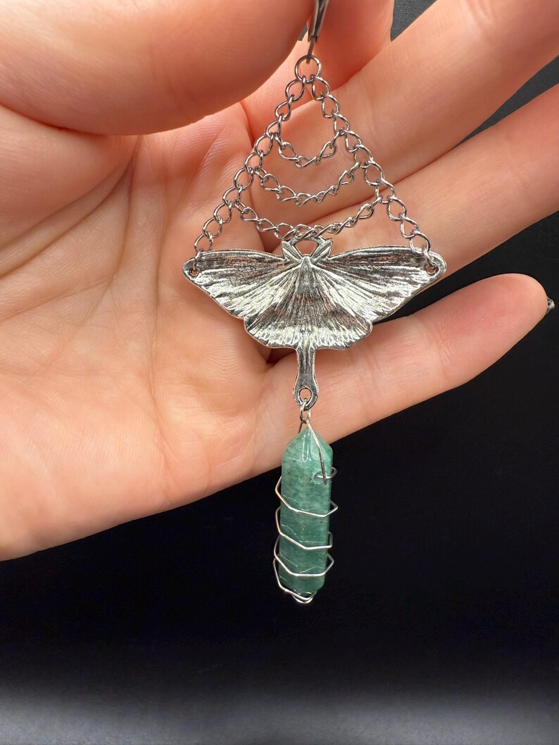 May include: A silver-toned pendant featuring a moth design with a green crystal. The moth has detailed wing patterns and hangs from a chain. The green crystal is wrapped in silver wire, creating a unique and elegant accessory.
