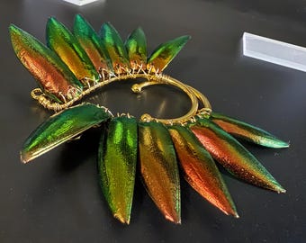May include: Ear cuff featuring iridescent green and copper-colored beetle wings. The wings are attached to a gold-colored wire frame and ear loop. The design is an artistic and unique piece of jewelry.