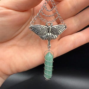 May include: Silver-toned pendant featuring a moth design with intricate wing details. The pendant hangs from a chain and connects to a green crystal wrapped in silver wire. The crystal is a light green color and is cylindrical in shape.