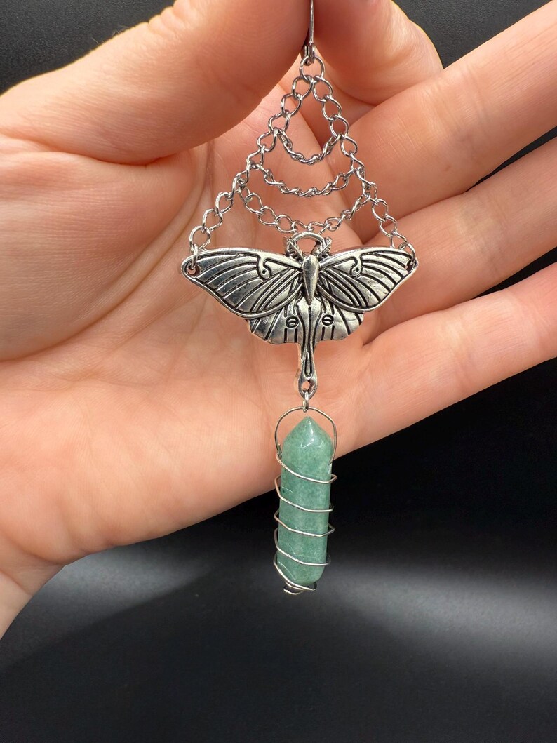 May include: Silver-toned pendant featuring a moth design with intricate wing details. The pendant hangs from a chain and connects to a green crystal wrapped in silver wire. The crystal is a light green color and is cylindrical in shape.