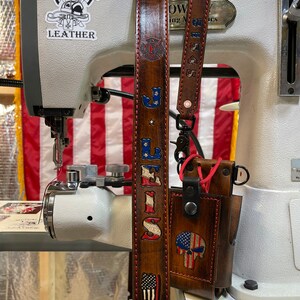 May include: Brown leather strap with red stitching, featuring the text "LEWIS" and an American flag design. A matching leather phone case with a skull and American flag design is also visible. The strap is near a sewing machine.