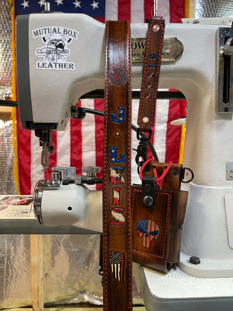 May include: Brown leather strap with red stitching, featuring the text "LEWIS" and an American flag design. A matching leather phone case with a skull and American flag design is also visible. The strap is near a sewing machine.
