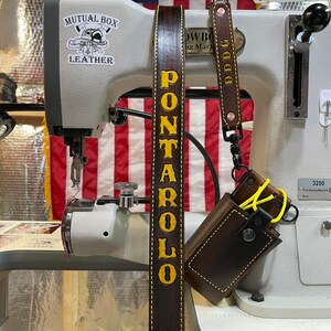 May include: A dark brown leather strap with the name "PONTAROLO" in yellow lettering. A smaller matching strap with "DDDG" is attached to a brown leather case. The background includes a sewing machine and an American flag.