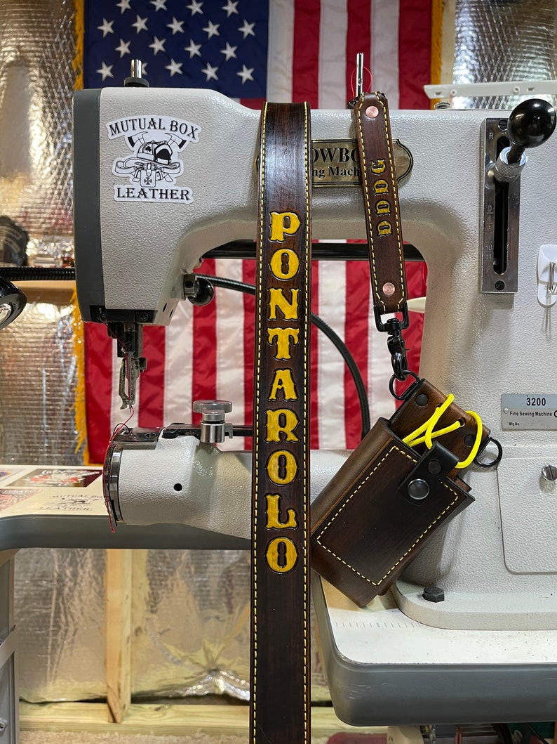 May include: A dark brown leather strap with the name "PONTAROLO" in yellow lettering. A smaller matching strap with "DDDG" is attached to a brown leather case. The background includes a sewing machine and an American flag.