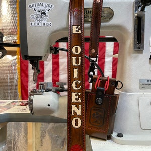 May include: A brown leather strap with the name "EQUICENO" in white letters. The strap has red stitching and a red emblem. A small brown leather pouch is attached. The background includes a sewing machine and an American flag.
