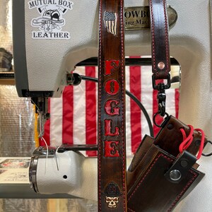 May include: A brown leather strap with the name "FOGLE" in red lettering and a small American flag design. The strap is attached to a sewing machine with a "Mutual Box Leather" sticker. A brown leather case with red stitching is also visible.
