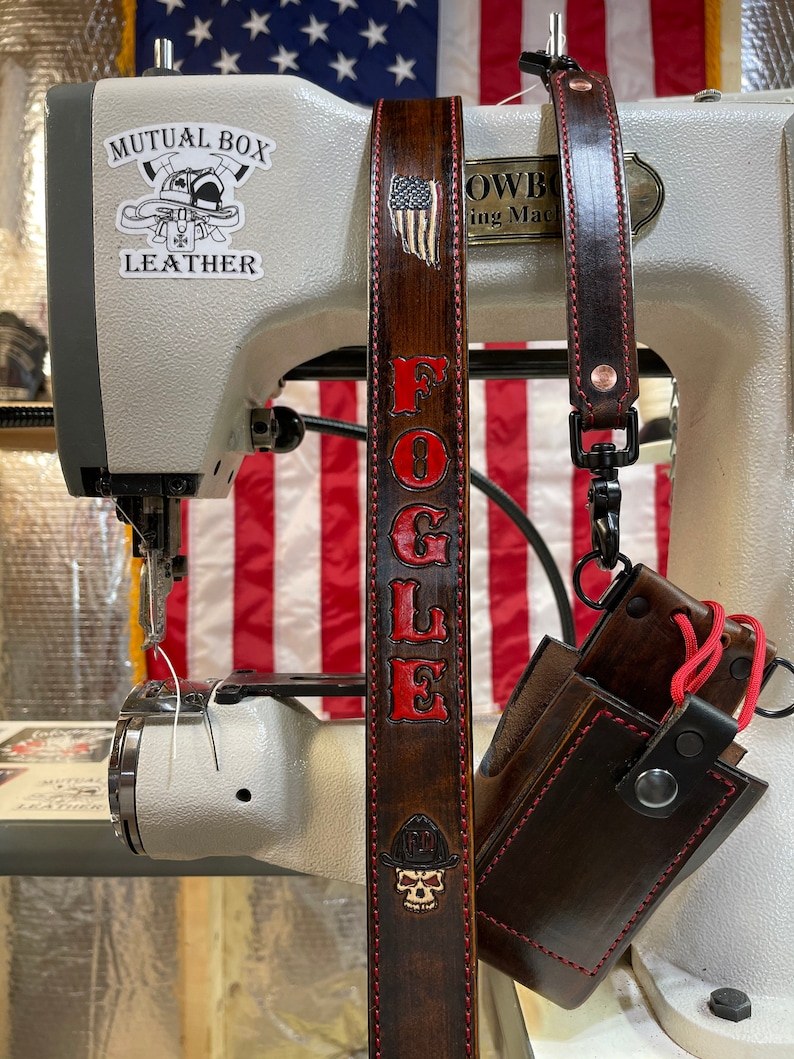 May include: A brown leather strap with the name "FOGLE" in red lettering and a small American flag design. The strap is attached to a sewing machine with a "Mutual Box Leather" sticker. A brown leather case with red stitching is also visible.