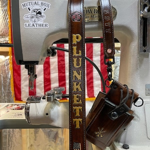 May include: Dark brown leather belt with the name "PLUNKETT" in gold lettering and an American flag design. The belt is hanging in front of a sewing machine with the words "Mutual Box Leather" on it.