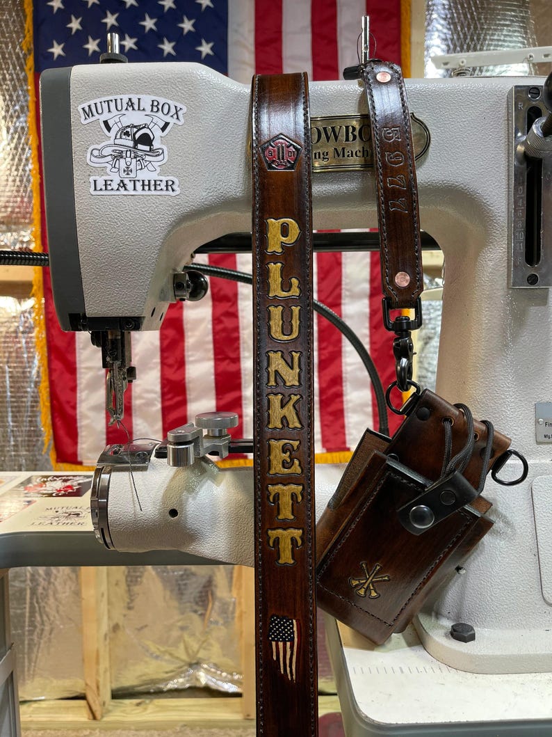 May include: Dark brown leather belt with the name "PLUNKETT" in gold lettering and an American flag design. The belt is hanging in front of a sewing machine with the words "Mutual Box Leather" on it.