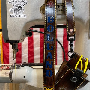 May include: A brown leather strap with the name "BOLAND" in blue lettering and a gold border. The strap also features a West Virginia University logo and a firefighter emblem. A small leather pouch is attached to the strap.