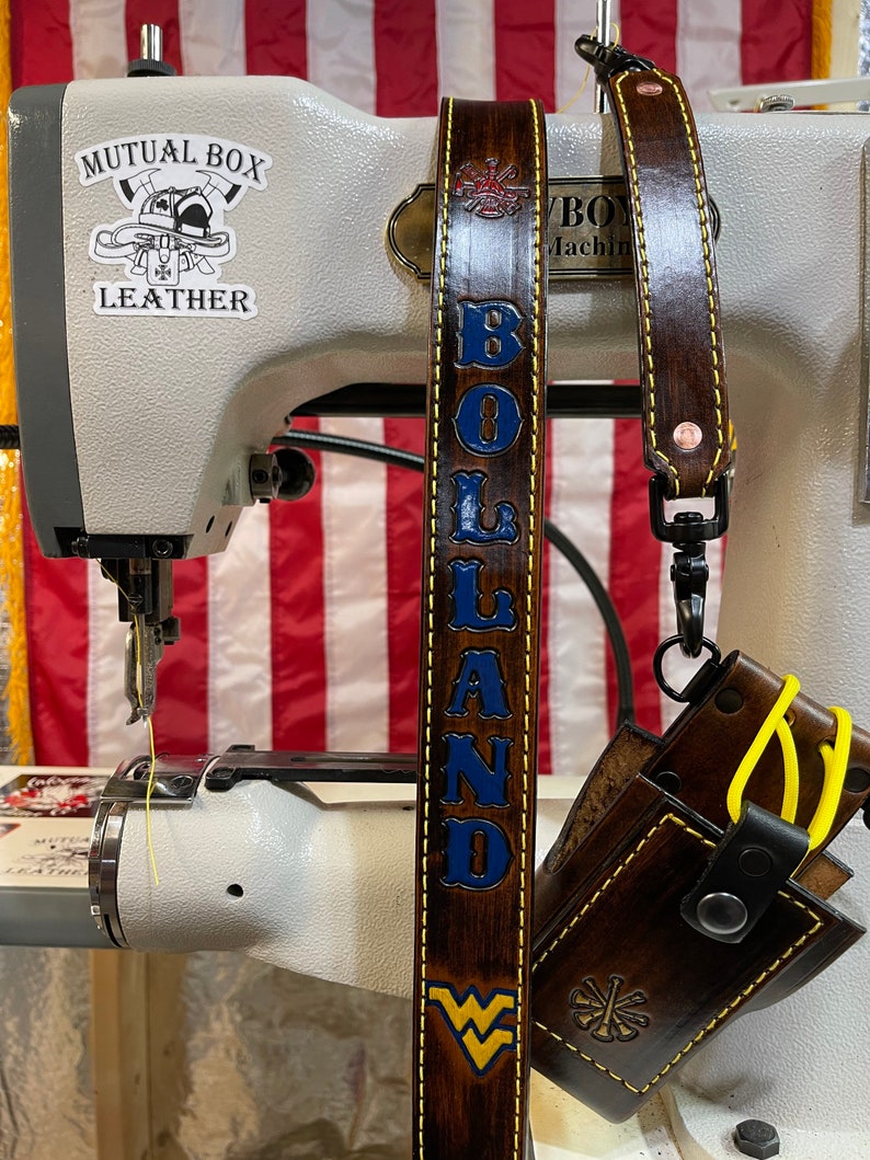 May include: A brown leather strap with the name "BOLAND" in blue lettering and a gold border. The strap also features a West Virginia University logo and a firefighter emblem. A small leather pouch is attached to the strap.