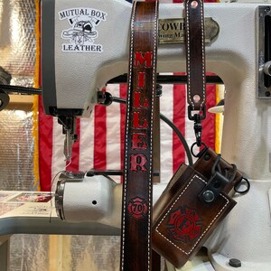 May include: Dark brown leather strap with the name "MILLER" in red lettering and white stitching. A matching leather case with a red firefighter emblem is attached. The background includes a sewing machine and an American flag.