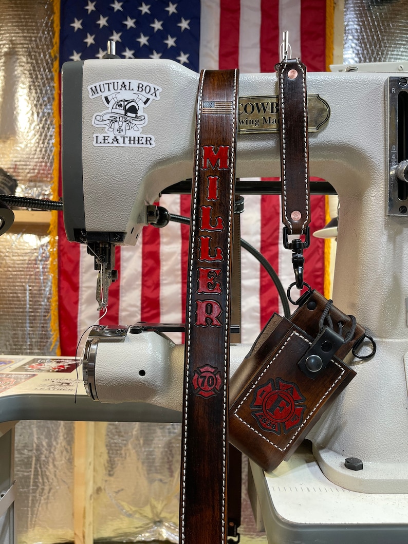 May include: Dark brown leather strap with the name "MILLER" in red lettering and white stitching. A matching leather case with a red firefighter emblem is attached. The background includes a sewing machine and an American flag.
