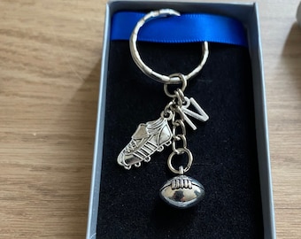 May include: A silver keychain with a football, a boot, and a charm with the letter 'V' hanging from a silver ring. The keychain is in a blue and silver gift box.